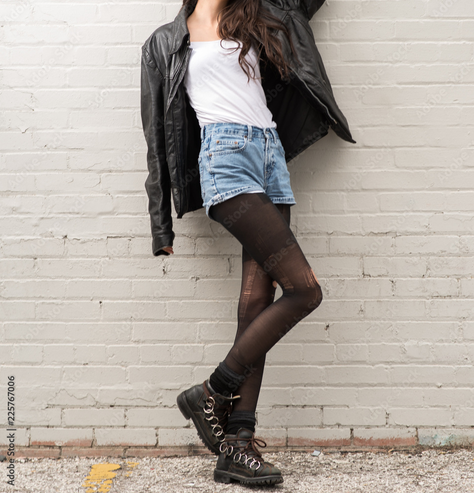 Young Woman in a Black Leather Jacket, Denim Shorts and Torn Stockings  Against a Brick wall Stock Photo | Adobe Stock