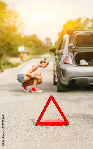 Young beautiful woman spection a rear tire on the side of the road. Car with problems and red triangle to warn other road users. photo