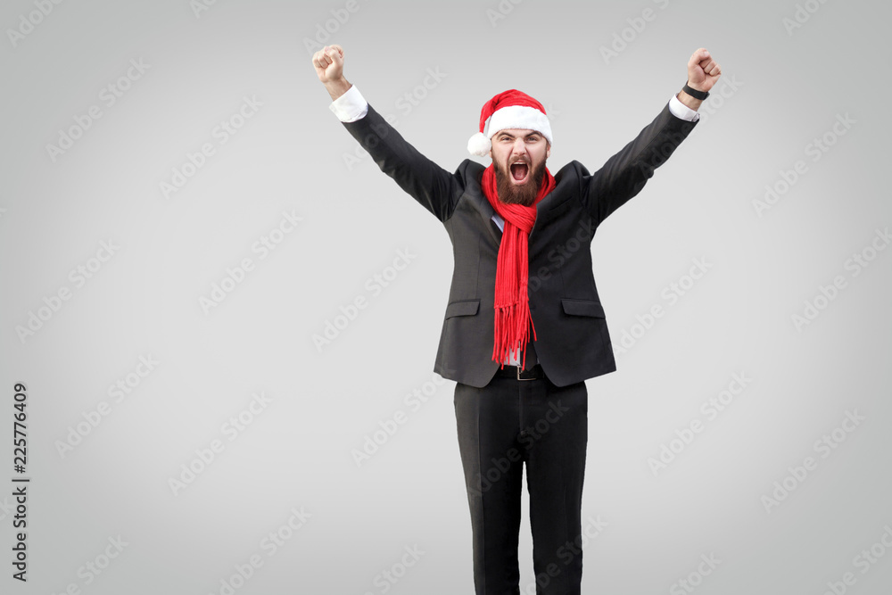 Bearded businessman in classic black suit, red scarf and new year hat, looking at camera with raised arms and screaming. Wining and success concept. Indoor studio shot, Isolated on gray background. Bearded businessman in classic black suit, red scarf and new year hat, looking at camera with raised arms and screaming. Wining and success concept. Indoor studio shot, Isolated on gray background.