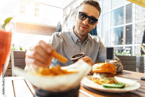 Close up of young attractive man eating french fries and burger at street cafe.