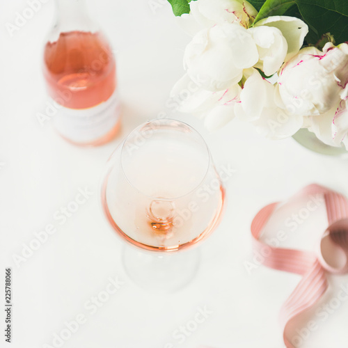 Rose wine in glass and bottle, pink decorative ribbon, peony flowers over white background, copy space, square crop. Summer celebration, wedding greeting card, invitation concept