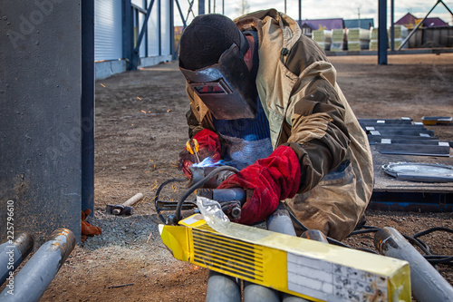 Welder weld metal pipe at construction site. A young man in brown uniform, welding mask and welders leathers, weld metal with a arc welding machine, blue sparks fly to the sides