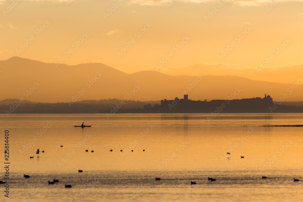 Beautiful view of Trasimeno lake (Umbria, Italy) at sunset, with orange tones, birds on water, a man on a canoe and Castiglione del Lago town on the background