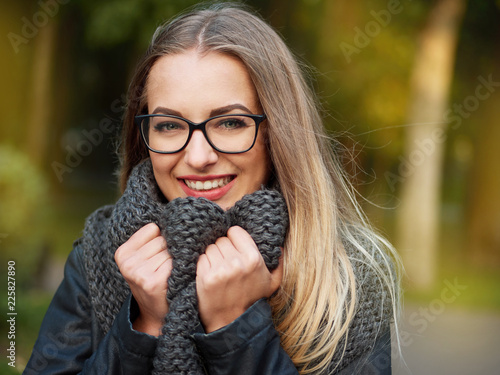 portrait of a beautiful stylish young blonde girl with make-up in black glasses leather jacket and knitted scarf freezes up smiles in the autumn cold park