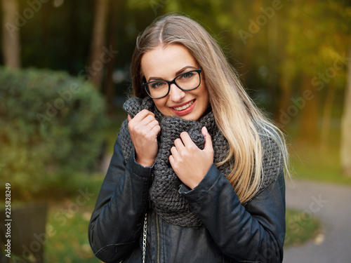 portrait of a beautiful stylish young blonde girl with make-up in black glasses leather jacket and knitted scarf freezes up smiles in the autumn cold park photo