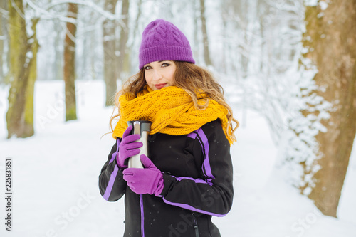 Young sporty woman in violet hat and plaid drinking hot coffee or tea after running morning exercise in winter snowy park.
