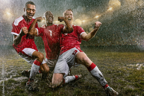 Soccer players on professional soccer night rain stadium. Three dirty players in rain drops emotionally rejoices victory. Men kneel and scream © Alex
