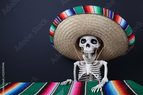 Day of the Dead.  human skeleton wearing a beautiful serape sombrero (hat) and sitting at a table covered with a colorful serape blanket