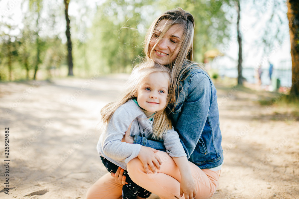 Happy smiling mother hugging her lovely little daughter outdoor. Lifestyle family. Adult cheerful female parent playing with her beautiful emotional child at nature in summer. Positive people faces.