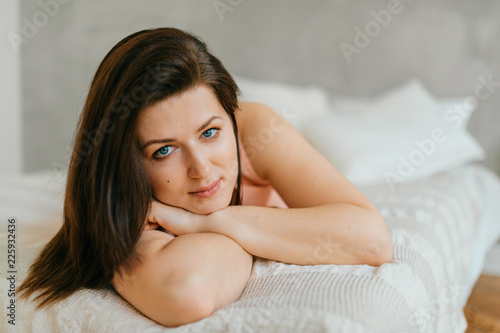 Beautiful brunette woman portrait luiyng on white bed at home in morning. Female after waking up. Cheerful adult married blue eyed girl in pink nighty with positive face smiling and relaxing on sofa