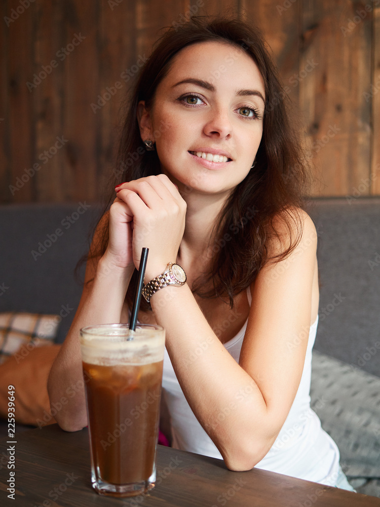 Beautiful cute brunette girl sitting in the cozy cafe near the window enjoying big cup coffee with straw and shy smiling Beautiful cute brunette girl sitting in the cozy cafe near the window enjoying big cup coffee with straw and shy smiling