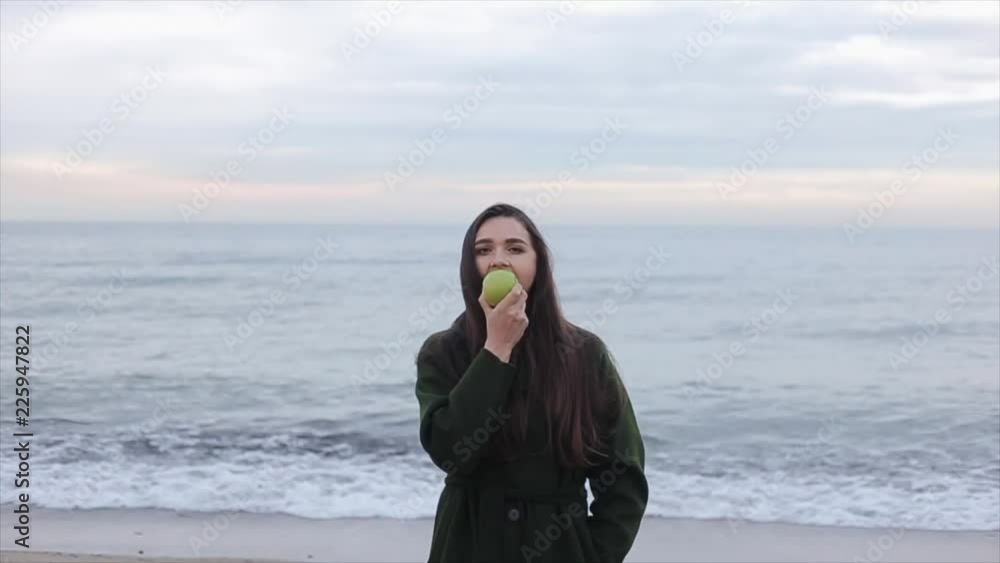 A portrait of a beautiful happy girl holding and eating an apple on the seashore on a cold day A portrait of a beautiful happy girl holding and eating an apple on the seashore on a cold day