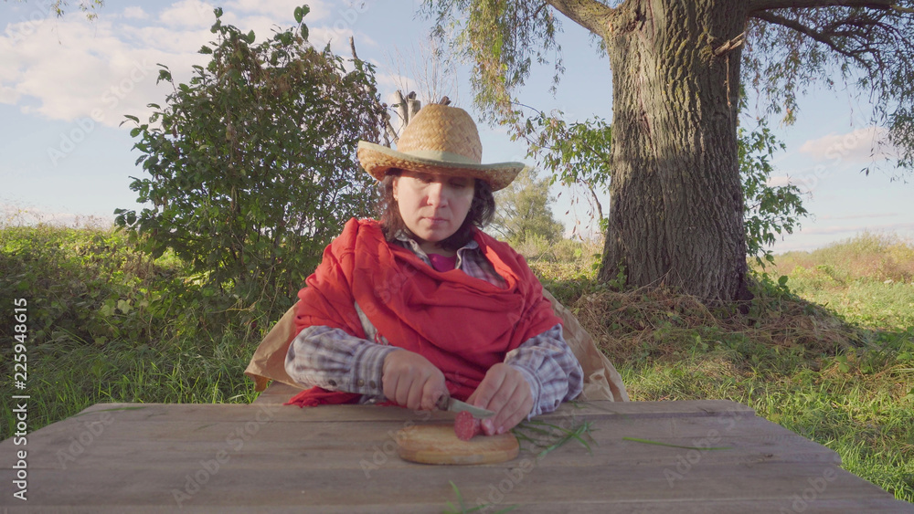 A young woman in a straw hat. Sits and cuts a piece of sausage. On the background of a large tree. Blue sky. Green grass and leaves.