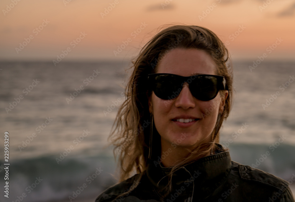 Beautiful blond curly hair woman wearing camouflaged jacket and sunglasses posing at sunset on the beach Beautiful blond curly hair woman wearing camouflaged jacket and sunglasses posing at sunset on the beach