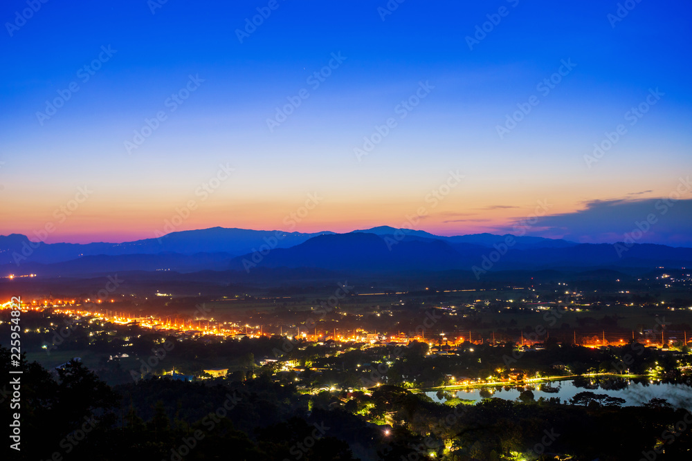 Aerial view at Doi Khao Kwai during blue zone after sunset. Mountain range as background with light tail on main road at Chiangrai city and big pond as foreground