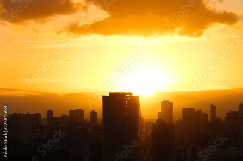 Pôr do sol com avião passando ao fundo, sobre os prédios de uma metrópole