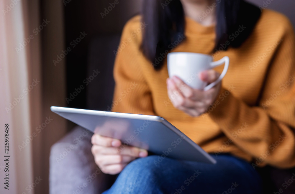 Reading Concept. Young Woman using Tablet and Drinking Coffee. Relaxing in Cozy House, Sit on the Chair, Front View and Selective Focus Reading Concept. Young Woman using Tablet and Drinking Coffee. Relaxing in Cozy House, Sit on the Chair, Front View and Selective Focus