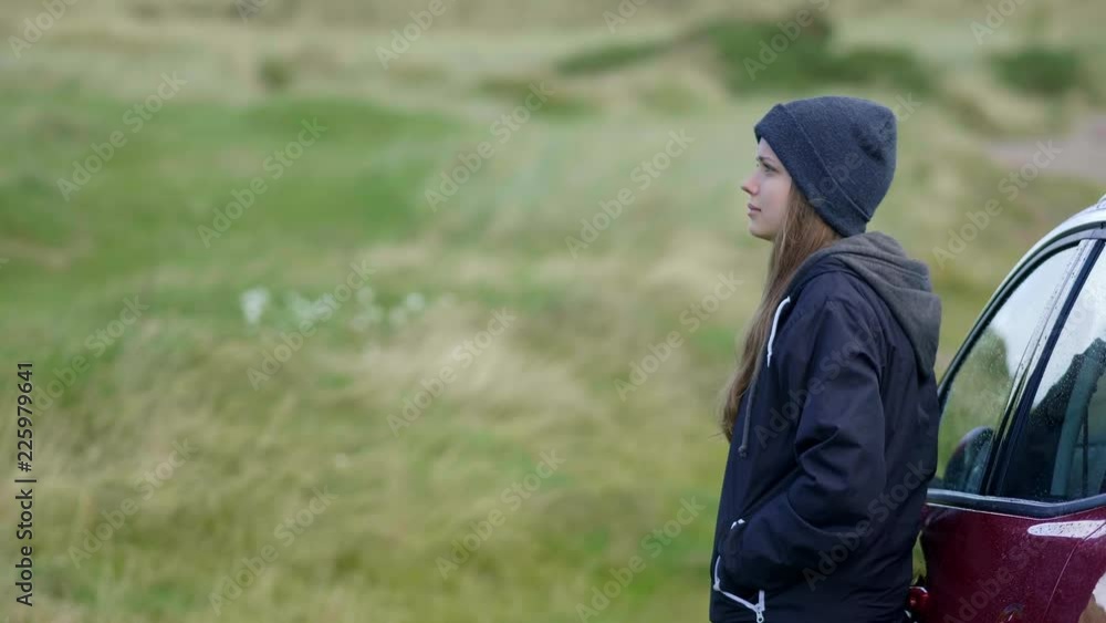 Young woman stands beside her car and looks over the green grassy fields Young woman stands beside her car and looks over the green grassy fields