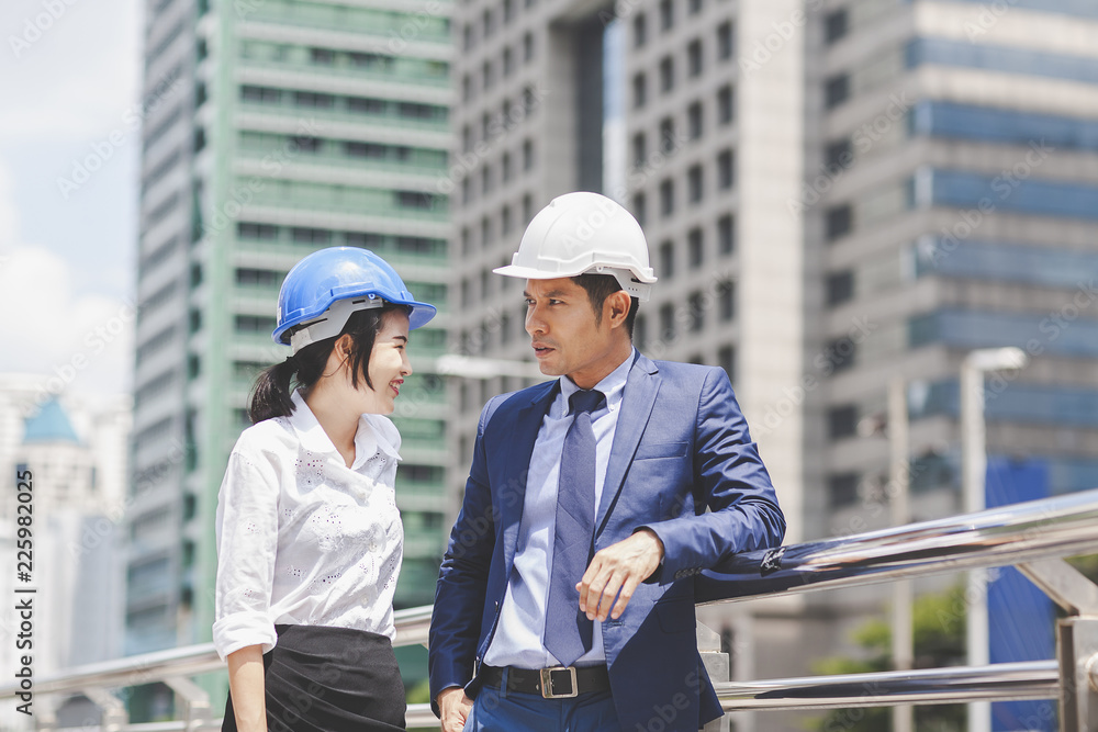 Two Asian engineers working or discussion on building site on modern city. Business people team in safety helmet talk New Project,Product Details with investment engineering and architecture concept