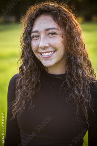 A beautiful Hispanic teen girl outdoor portrait. Cute and smiling woman with brown hair looking at the camera standing in a scenic field outdoors