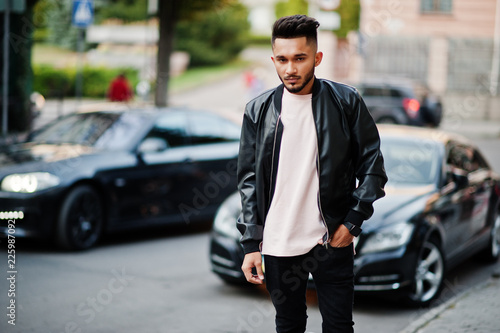 Stylish indian beard man at black leather jacket against car. India model posed at streets of city.