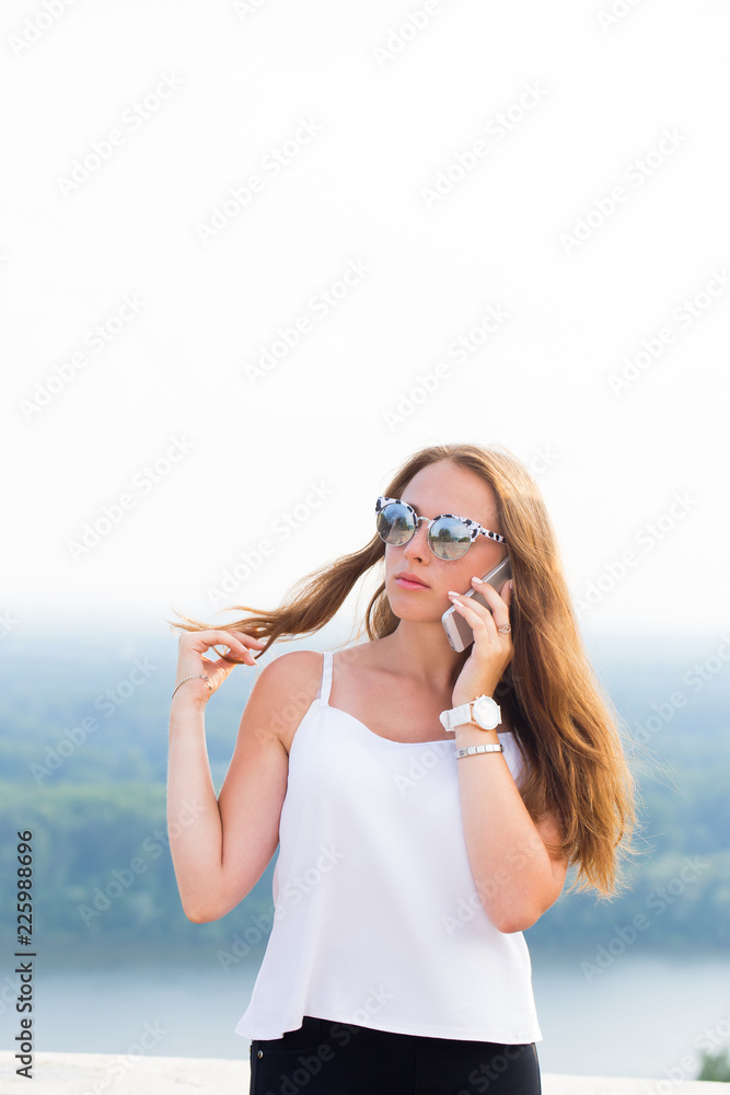 Attractive young woman with long curly hair, in sunglasses, white top and black pants talking on the phone
