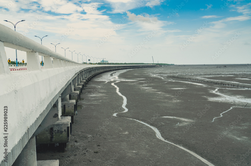 Curved coastal concrete highway road with lower water in the mud beach with blue sky and clouds. Road trip travel. Summer journey at tropical Cement bunting bridge. Nature abstract background.