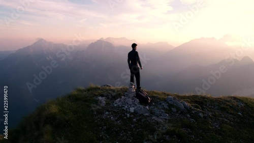 Aerial - Flying around young man standing victoriously on top of the mountain at sunset