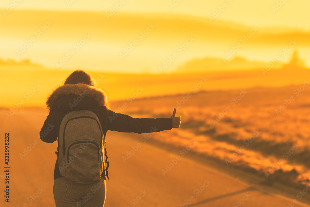 Young woman in the winter jacket with a backpack on the road against the background of the sunrise autumn field. Travel and tourism concept