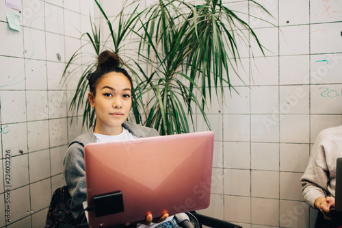 Portrait of confident young female hacker using laptop while sitting against tile wall at small creative office photo