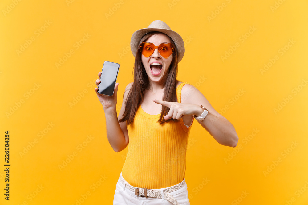 Portrait of excited amazed woman in summer hat, orange glasses keep mouth wide open, look surprised, hold mobile phone with blank empty screen isolated on yellow background. People emotions concept. Portrait of excited amazed woman in summer hat, orange glasses keep mouth wide open, look surprised, hold mobile phone with blank empty screen isolated on yellow background. People emotions concept.