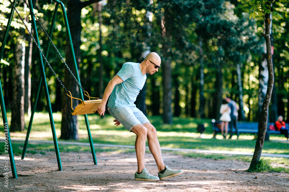 Fototapeta premium Odd adult bald man in sunglasses jumping from swing on ground in motion. Childhood behavior. Hulking awkward foolish comic male have fun on children playground outdoor in city park in summer day.