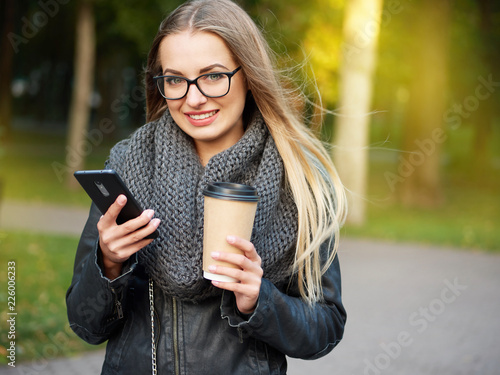 Portrait of a beautiful stylish young blonde with make-up in black leather jacket glasses and a knitted scarf. Frozen girl drinks hot drink and talks on the phone in the autumn cold park