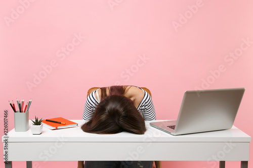 Young frustrated exhausted woman laid her head down on the table sit work at white desk with contemporary pc laptop isolated on pastel pink background. Achievement business career concept. Copy space. photo