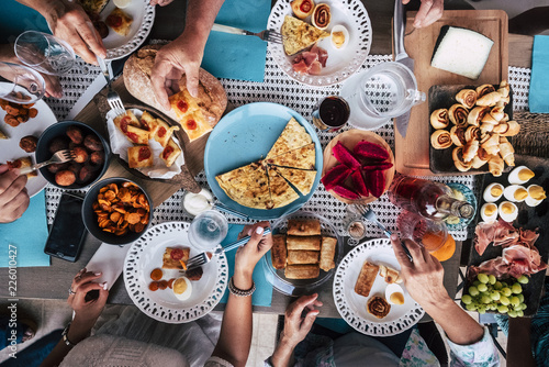 Above aerial view of group of friends having fun eating together at lunch or dinner with a table full of different and colorful food and technology mobile phone. mix of hands of caucasian people 
