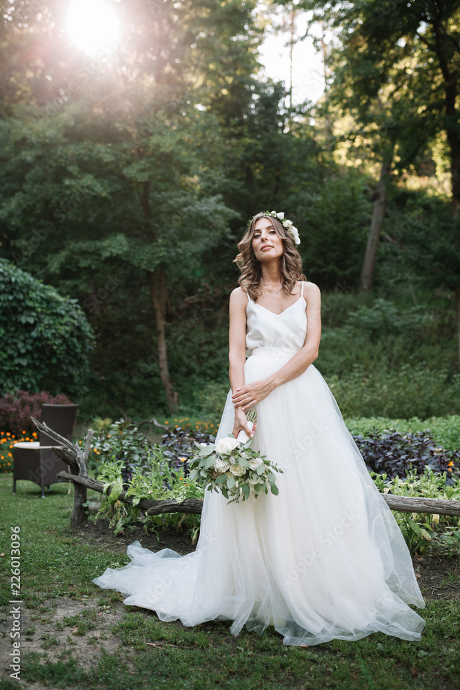 Beautiful blonde happy bride in elegant white dress in a wreath with bouquet outdoors