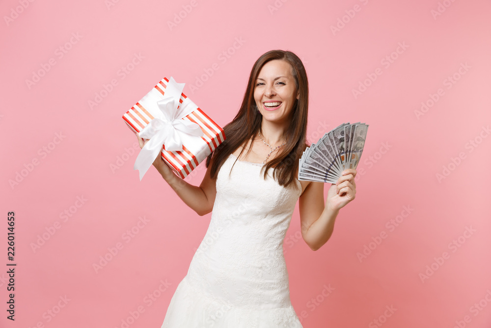 Portrait of laughing bride woman in white wedding dress hold bundle lots of dollars, cash money, red box with gift, present isolated on pink pastel background. Wedding celebration concept. Copy space. Portrait of laughing bride woman in white wedding dress hold bundle lots of dollars, cash money, red box with gift, present isolated on pink pastel background. Wedding celebration concept. Copy space.