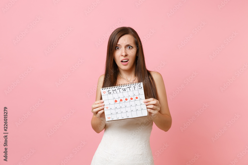 Shocked concerned bride woman in white wedding dress holding female periods calendar for checking menstruation days isolated on pink background. Medical, healthcare, gynecological concept. Copy space. Shocked concerned bride woman in white wedding dress holding female periods calendar for checking menstruation days isolated on pink background. Medical, healthcare, gynecological concept. Copy space.