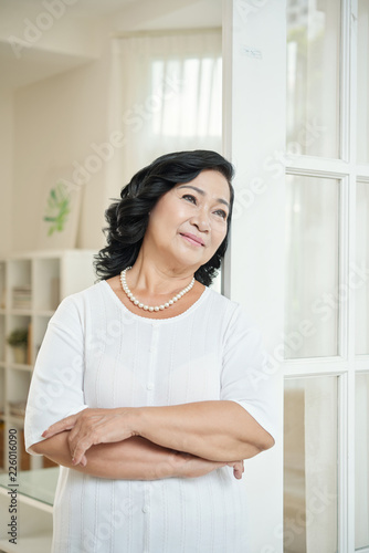 Good-looking elderly Asian woman standing in apartment, leaning on doors, looking away thoughtfully and smiling