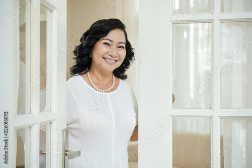 Attractive elegant mature Asian woman standing near doorway in her apartment and smiling at camera happily