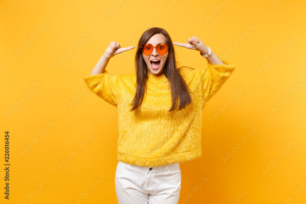 Excited happy young woman in fur sweater and heart orange glasses pointing index fingers on head isolated on bright yellow background. People sincere emotions, lifestyle concept. Advertising area. Excited happy young woman in fur sweater and heart orange glasses pointing index fingers on head isolated on bright yellow background. People sincere emotions, lifestyle concept. Advertising area.