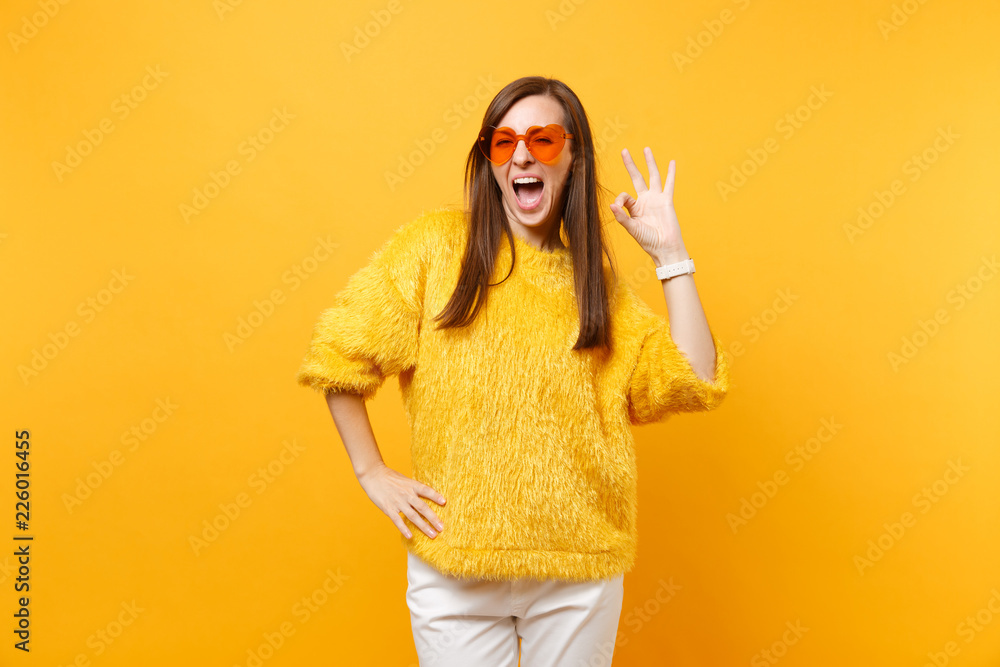 Portrait of joyful young woman in fur sweater, white pants and heart orange glasses showing OK sign isolated on bright yellow background. People sincere emotions, lifestyle concept. Advertising area. Portrait of joyful young woman in fur sweater, white pants and heart orange glasses showing OK sign isolated on bright yellow background. People sincere emotions, lifestyle concept. Advertising area.