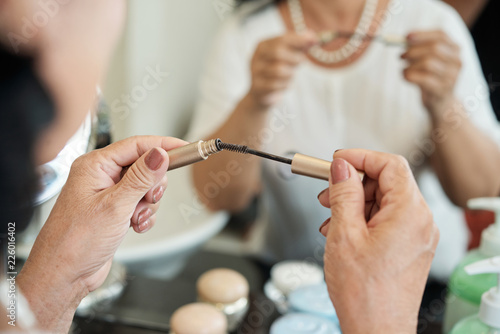 Close-up view of unrecognizable aged woman holding mascara brush in front of mirror in bathroom