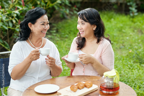 Beautiful stylish mature Asian women chatting and smiling happily while sitting at table with fresh tea and croissants outdoors