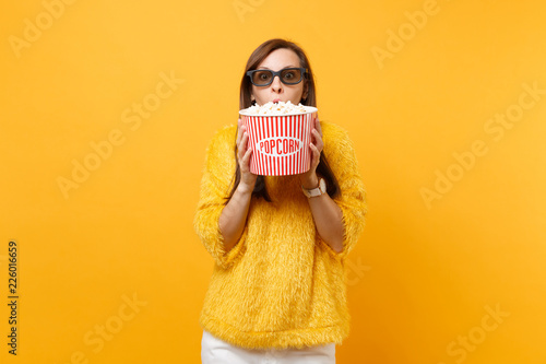 Portrait of shocked young girl in 3d imax glasses watching movie film, covering mouth with bucket of popcorn isolated on bright yellow background. People sincere emotions in cinema, lifestyle concept. photo