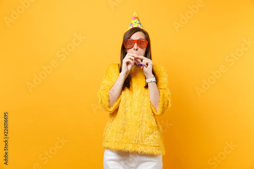 Portrait of Amazed young woman in orange funny eyeglasses, birthday party hat with playing pipe celebrating isolated on bright yellow background. People sincere emotions lifestyle. Advertising area. © ViDi Studio