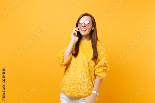 Cheerful young woman in fur sweater, heart glasses talking on mobile phone conducting pleasant conversation isolated on bright yellow background. People sincere emotions, lifestyle. Advertising area.