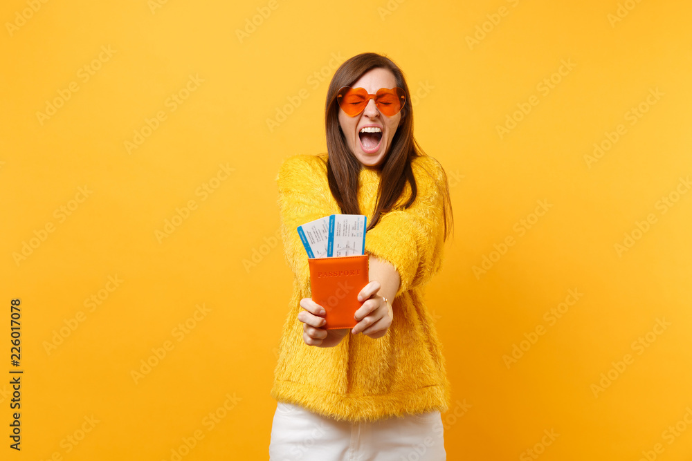 Crazy pretty young woman with closed eyes in orange heart glasses screaming, holding passport boarding pass tickets isolated on yellow background. People sincere emotions, lifestyle. Advertising area. - obrazy, fototapety, plakaty 