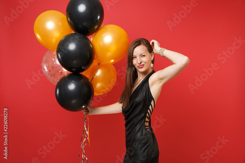 Cheerful young woman in black dress celebrating keeping hand near head holding air balloons isolated on bright red background. St. Valentine's Day Happy New Year birthday mockup holiday party concept. © ViDi Studio