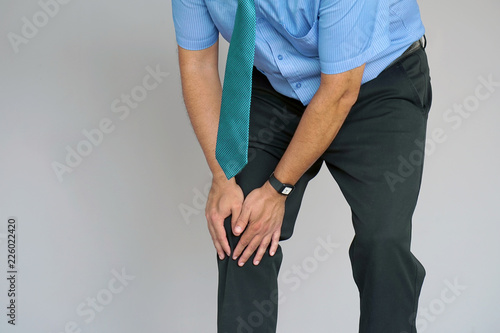 Pain In Knee. Close-up Businessman Leg With Painful Kneeson on gray background. Man Feeling Joint Pain, Having Health Issues And Touching Leg With Hands. Body And Health Care Concept.